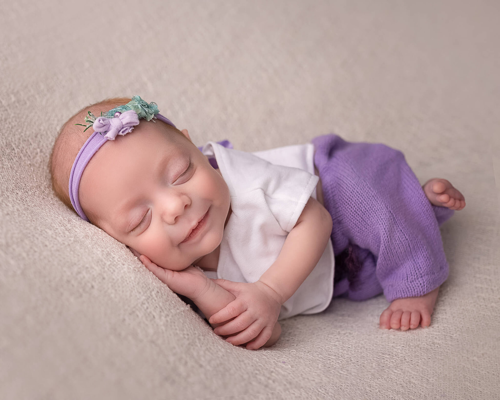 newborn in purple and white outfit sleeping with hand under cheek and smiling during newborn photography session