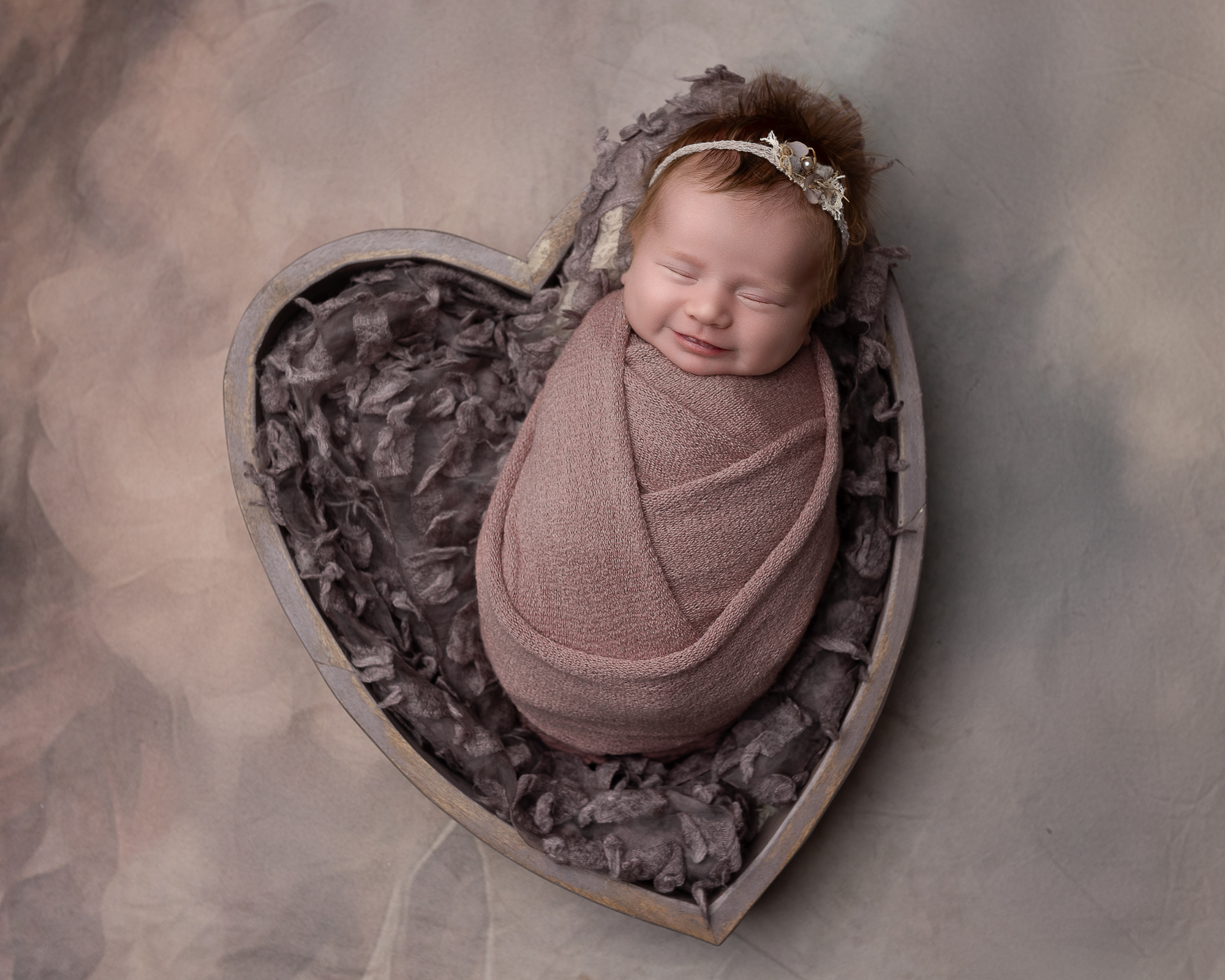 Akron newborn photography portrait of a smiling newborn wrapped in soft mauve tones and posed in a heart-shaped bowl
