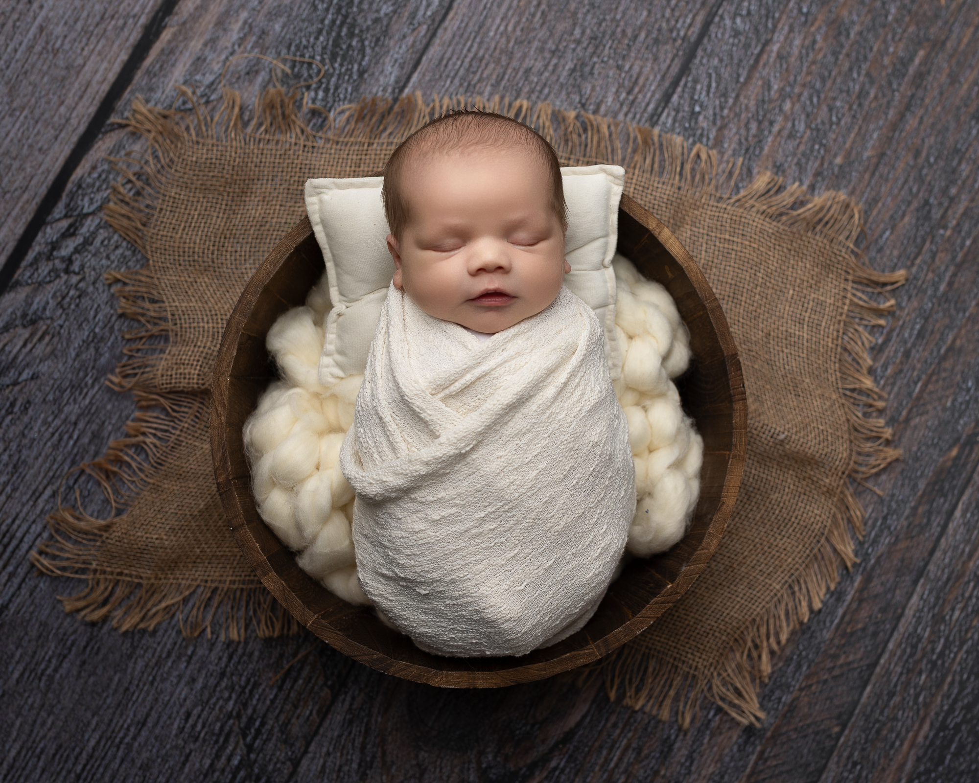Sleeping newborn wrapped in a soft white blanket and posed in a wooden bowl during studio photo session prior to visiting akron pediatrician