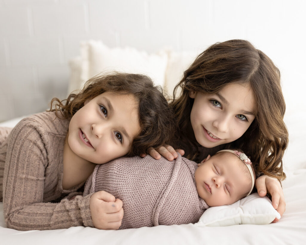 Older sisters lying beside their newborn baby sister during a newborn photography session wearing soft neutral outfits